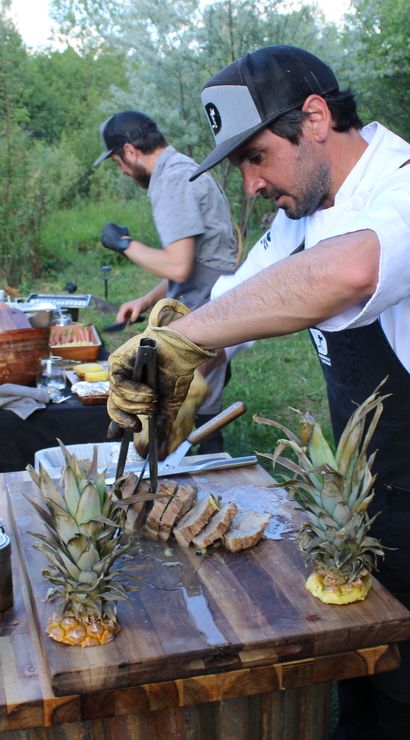 Chef in apron slicing grilled meat on a wooden cutting board with pineapple garnishes at a rustic outdoor garden barbecue prep station.