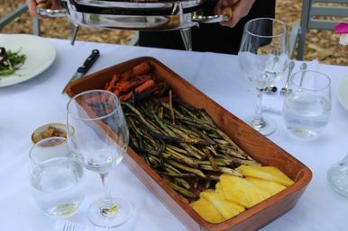 Al fresco table setting with a wooden tray of charred green beans, roasted baby carrots and pineapple slices next to wine and water glasses on a white tablecloth.