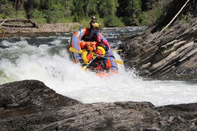Thrilling whitewater rafting scene: group of paddlers in helmets and bright life jackets navigating a rocky mountain river rapid in an orange inflatable raft.
