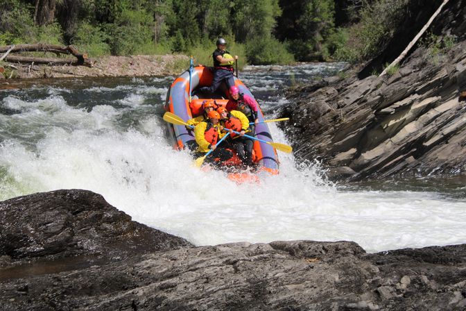 Thrilling whitewater rafting scene: group of paddlers in helmets and bright life jackets navigating a rocky mountain river rapid in an orange inflatable raft.