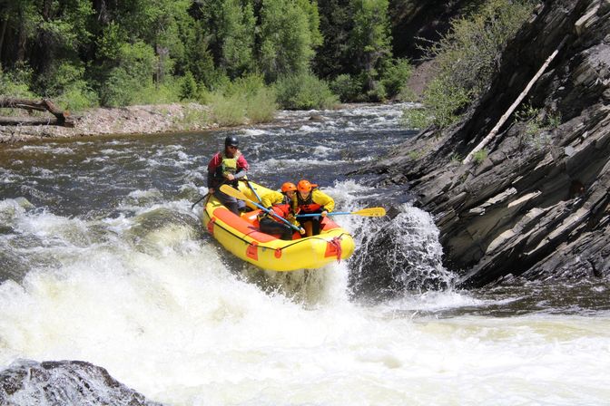 Whitewater rafting adventure: a yellow inflatable raft with two paddlers in orange helmets and a guide riding a steep river rapid, splashing through whitewater against a rocky canyon and forest backdrop.