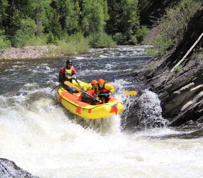 Whitewater rafting adventure: a yellow inflatable raft with two paddlers in orange helmets and a guide riding a steep river rapid, splashing through whitewater against a rocky canyon and forest backdrop.