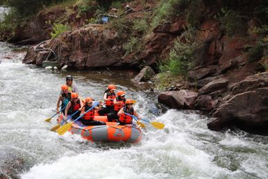Group of rafters in orange helmets and life jackets paddling an orange inflatable through whitewater rapids in a rocky mountain river canyon.