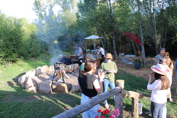 Friends enjoying an outdoor garden barbecue around a stone fire pit and tripod grill in a wooded backyard setting with picnic tables, umbrellas and colorful flowers.