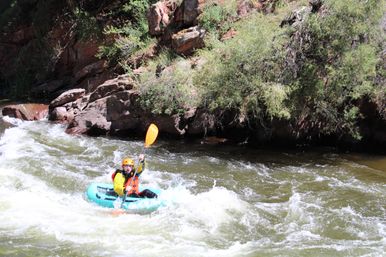 Solo kayaker in a bright helmet and life vest paddling a teal inflatable kayak through whitewater rapids beside a rocky, shrub-lined riverbank