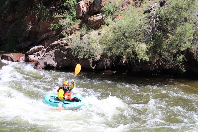 Solo kayaker in a bright helmet and life vest paddling a teal inflatable kayak through whitewater rapids beside a rocky, shrub-lined riverbank