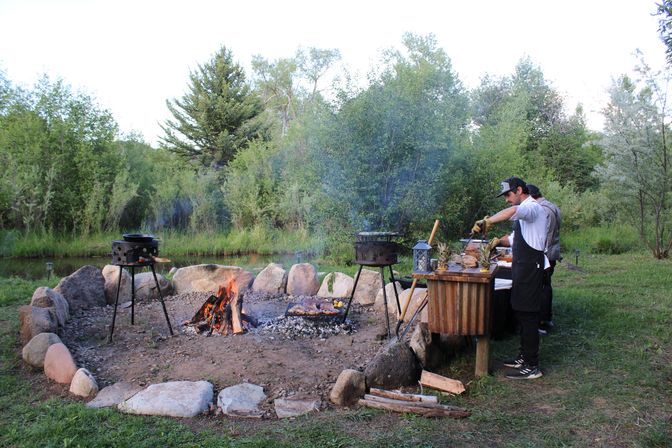 Lakeside outdoor cookout with two people grilling on tripod grills over an open campfire inside a stone fire ring, prepping food at a rustic wooden table by a pond and green trees.