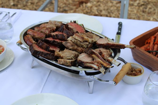 Juicy grilled lamb chops and sliced medium-rare steak on a stainless serving platter atop a white tablecloth for outdoor dining, with roasted carrots and a small dipping sauce nearby.