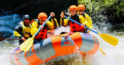 Four-person whitewater rafting team in orange helmets and life jackets paddling an orange inflatable raft through splashing rapids on a sunny mountain river