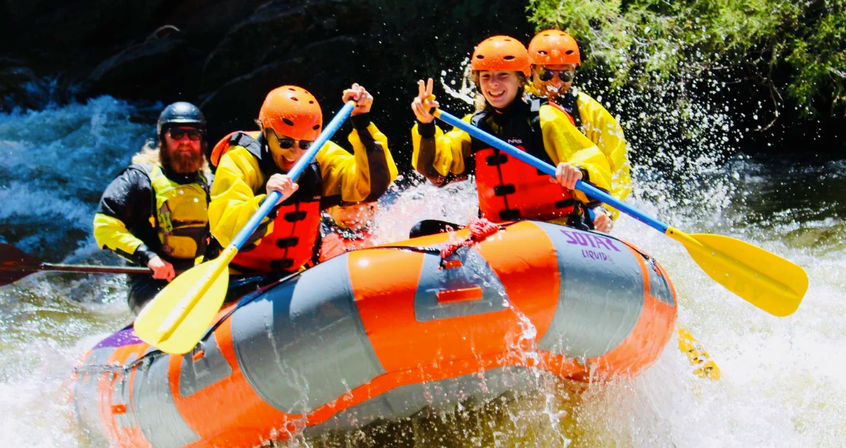 Four-person whitewater rafting team in orange helmets and life jackets paddling an orange inflatable raft through splashing rapids on a sunny mountain river