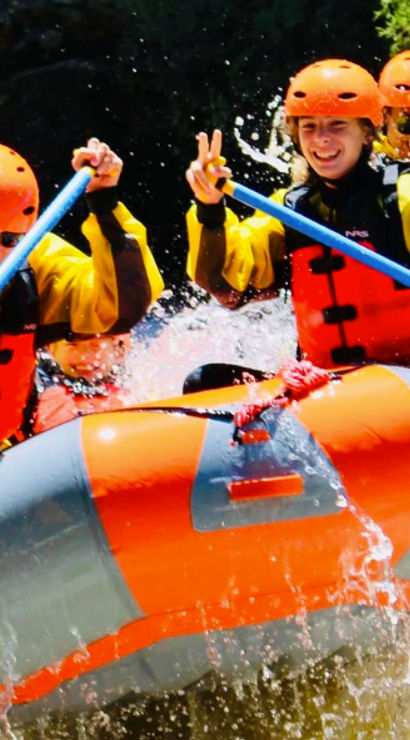 Four-person whitewater rafting team in orange helmets and life jackets paddling an orange inflatable raft through splashing rapids on a sunny mountain river