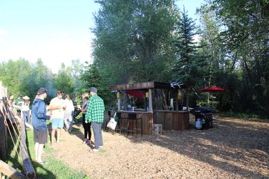 Outdoor summer gathering at a rustic wooden bar in a wooded backyard, people chatting near a grill and red umbrella on wood-chip ground.