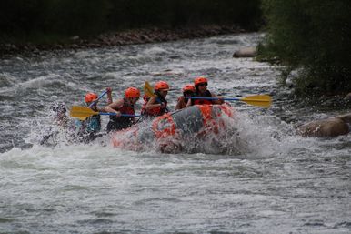 Whitewater rafting group in orange helmets and life jackets paddling an orange inflatable raft through churning rapids on a tree-lined river, water splashing around them.