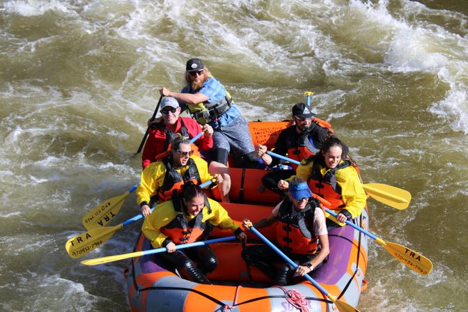 Six rafters in an orange inflatable raft wearing bright life jackets and using yellow paddles navigate choppy whitewater rapids on a sunny river rafting adventure.