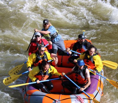 Six rafters in an orange inflatable raft wearing bright life jackets and using yellow paddles navigate choppy whitewater rapids on a sunny river rafting adventure.