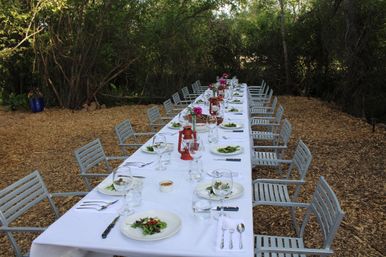 Long al fresco dining table in a wooded garden, set with a white tablecloth, salad plates and wine glasses, red lantern centerpieces and metal chairs on a wood‑chip floor.