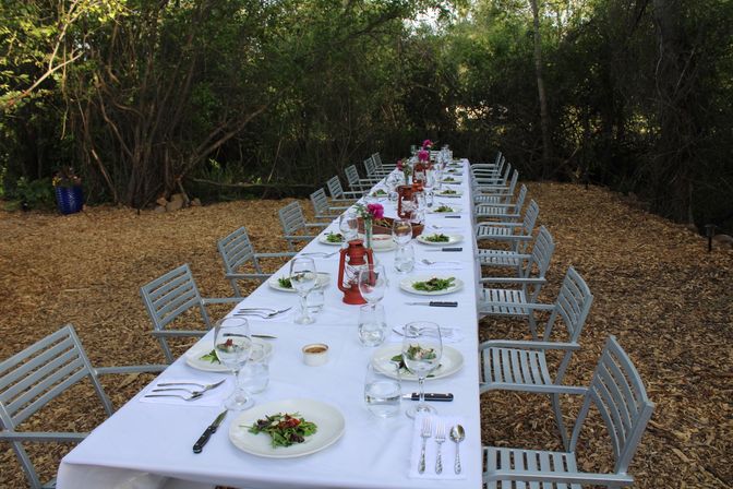 Long al fresco dining table in a wooded garden, set with a white tablecloth, salad plates and wine glasses, red lantern centerpieces and metal chairs on a wood‑chip floor.