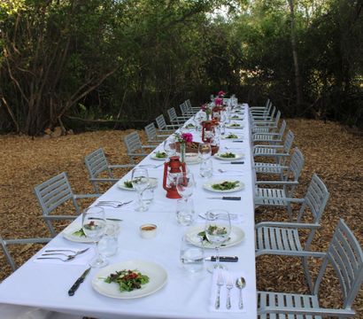 Long al fresco dining table in a wooded garden, set with a white tablecloth, salad plates and wine glasses, red lantern centerpieces and metal chairs on a wood‑chip floor.