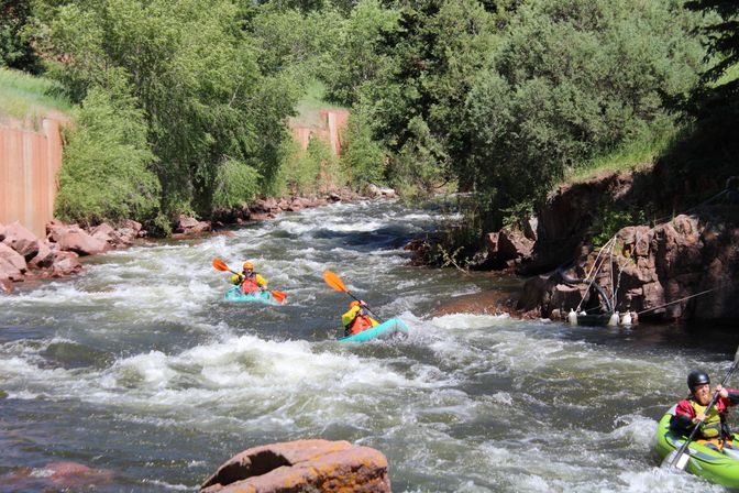 Three kayakers in colorful kayaks navigating whitewater rapids on a rocky, tree-lined mountain river, splashing through a sunny outdoor adventure