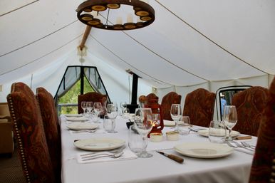 Elegant outdoor dining in a white canvas glamping tent: long table with white linen, plates, wine glasses, cutlery, lantern centerpiece and paisley-upholstered chairs under a rustic chandelier