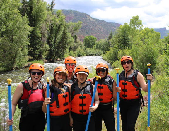 Six rafters in orange helmets and life jackets holding blue paddles smiling by a fast mountain river surrounded by green trees and hills