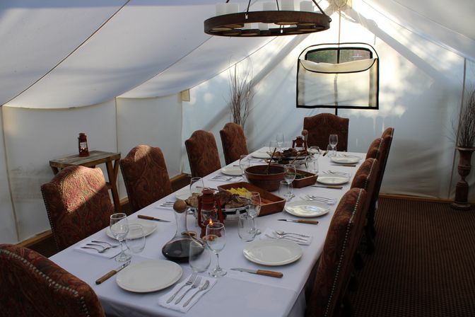 Cozy glamping dining setup: long white-tablecloth table with patterned upholstered chairs, wine glasses, wooden serving bowls and lantern centerpiece inside a white canvas tent.