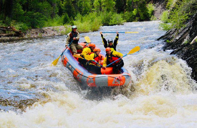 Group of rafters in an orange inflatable raft paddling through whitewater rapids on a forested mountain river, wearing helmets and life jackets, one rafter cheering with paddle raised