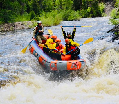 Group of rafters in an orange inflatable raft paddling through whitewater rapids on a forested mountain river, wearing helmets and life jackets, one rafter cheering with paddle raised