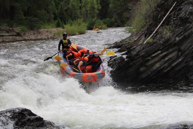Thrilling whitewater rafting scene: group wearing orange helmets paddling an inflatable raft through frothy rapids in a forested mountain river gorge.