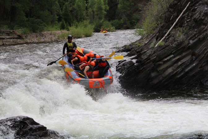 Thrilling whitewater rafting scene: group wearing orange helmets paddling an inflatable raft through frothy rapids in a forested mountain river gorge.