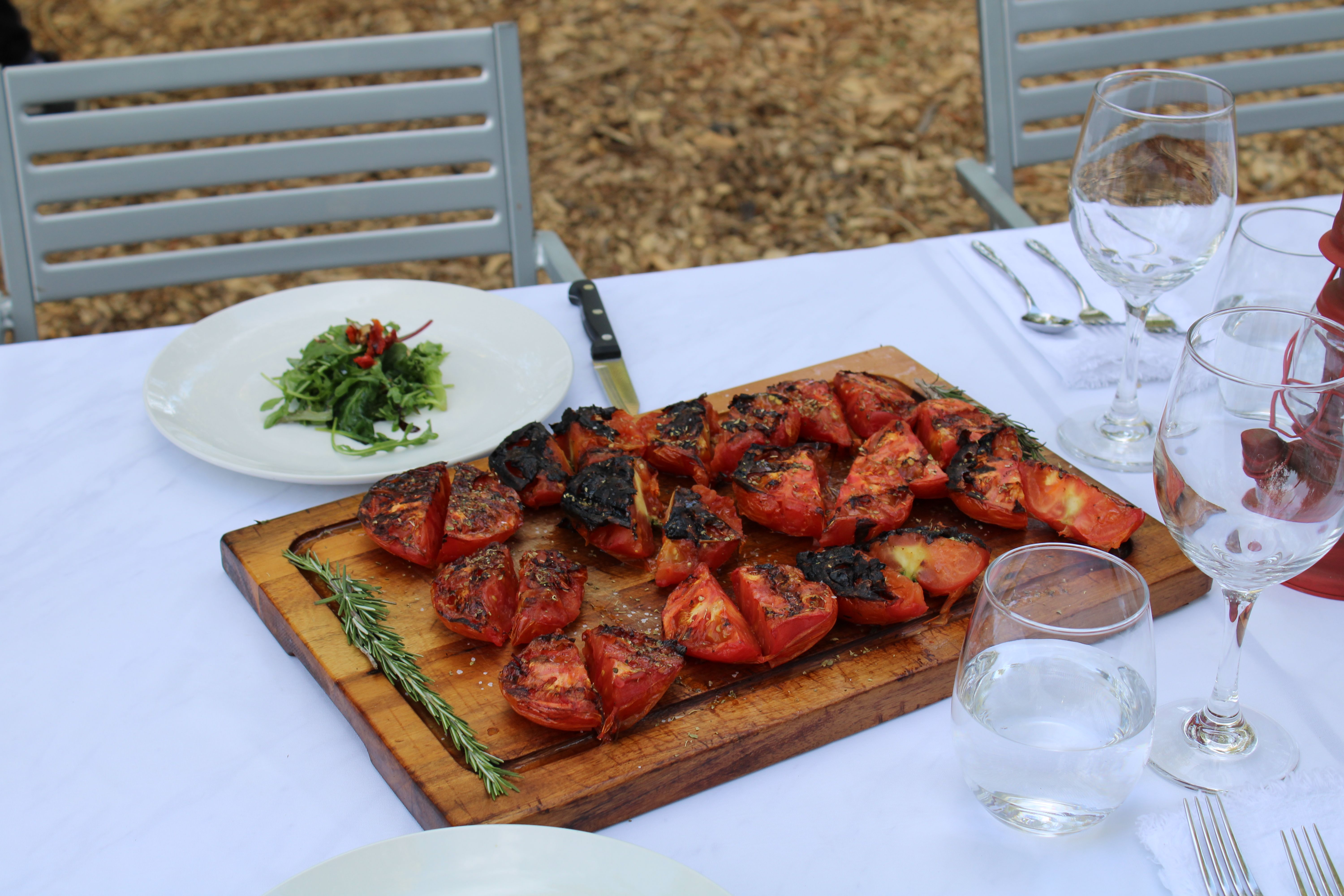 Charred roasted tomato halves on a wooden cutting board garnished with rosemary, set on a white-clothed outdoor dining table with wine glasses and a small green salad