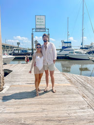 Smiling couple in casual summer outfits on a wooden marina dock — woman wearing heart-shaped sunglasses — sailboats moored in calm water under a bright blue sky.