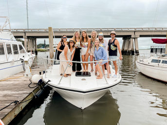 Nine friends posing on the bow of a white motorboat at a marina dock, smiling under a cloudy sky with a concrete bridge and calm water in the background.