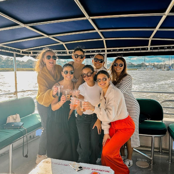 Seven friends on a covered party boat toasting drinks, wearing sunglasses and casual outfits, with a sunny marina and sailboats along the waterfront in the background.