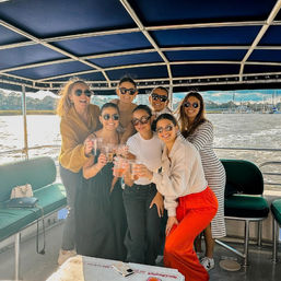 Seven friends on a covered party boat toasting drinks, wearing sunglasses and casual outfits, with a sunny marina and sailboats along the waterfront in the background.