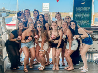 Group of women on a sunny marina dock posing for a girls trip photo, wearing swimsuits and heart-shaped sunglasses while holding sparkly disco-ball cups with boats and water in the background.