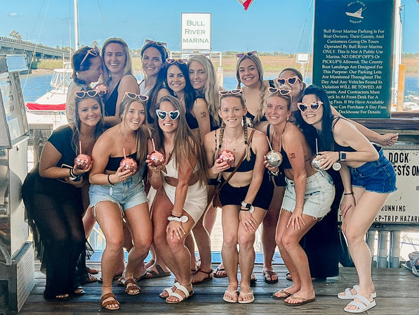 Group of women on a sunny marina dock posing for a girls trip photo, wearing swimsuits and heart-shaped sunglasses while holding sparkly disco-ball cups with boats and water in the background.