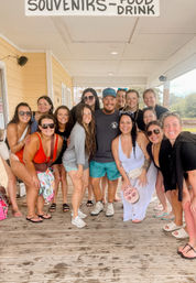 Smiling group of beach friends posing on a coastal wooden boardwalk under a 'SOUVENIRS - FOOD - DRINK' sign, wearing swimsuits and summer clothes beside a pale yellow building.