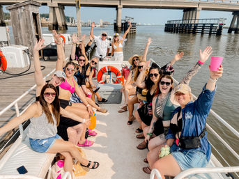 Smiling group of friends on a party boat at a marina under a bridge, waving and holding drinks on a sunny waterfront day