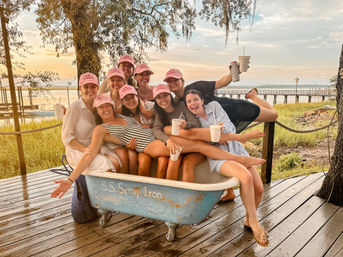 Group of smiling friends in matching pink caps packed into a vintage clawfoot bathtub on a waterfront wooden deck, holding drinks with a grassy marsh, pier, and sunset sky in the background.