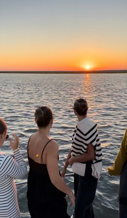 Group of people on a boat deck watching a golden sunset over calm water, sun reflecting on ripples; two wearing striped sweaters and hair in buns.