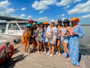 Group of ten women in colorful cowboy hats posing on a wooden marina dock beside a white boat, holding oversized face cutouts and drinks under a sunny blue sky with a bridge in the background.