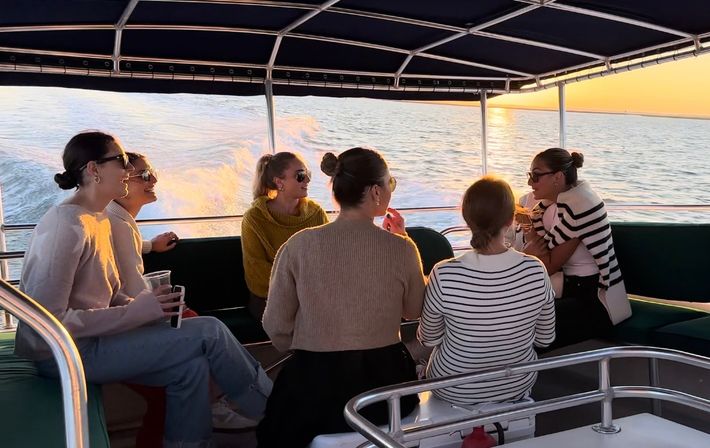 Sunset boat cruise on calm ocean: six friends in sweaters and sunglasses chat and sip drinks under a covered deck as the motorboat leaves a glowing wake.