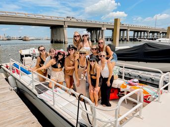 Group of smiling women in swimsuits and summer hats posing on a pontoon boat at a sunny marina dock with a concrete bridge and boats in the background.