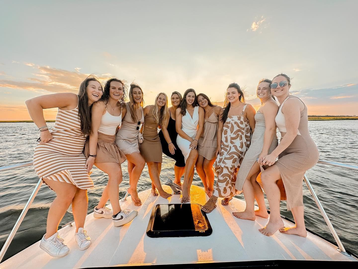 Ten women laughing and posing on the bow of a boat during a golden coastal sunset, wearing summer dresses and casual shoes with calm water and sky in the background.