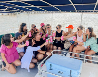 Group of friends cheering with pink canned drinks on a covered pontoon boat over calm coastal waters — summer boat party vibe