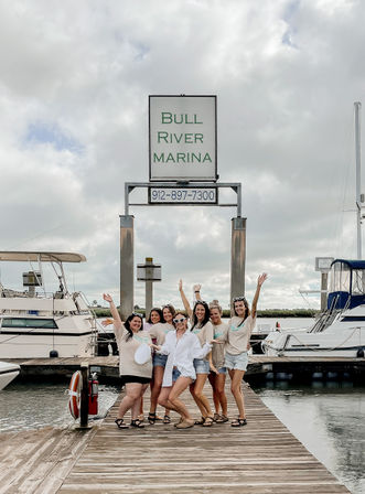 Group of friends cheering on a wooden marina dock beside moored yachts under a cloudy sky, waterfront coastal scene with an entrance sign in the background.