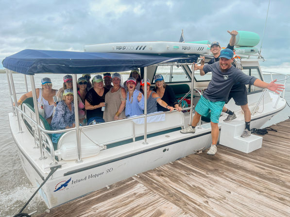 Smiling group of adults wearing colorful caps aboard a covered white passenger boat tied to a wooden dock, two men playfully posing on the pier under a cloudy sky over calm coastal water.
