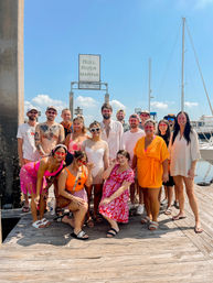 Group of friends posing on a sunny marina dock with sailboats and blue sky, colorful summer outfits and sunglasses, waterfront celebration vibe