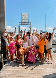 Energetic group celebrating on a sunny wooden dock at a coastal marina, colorful summer outfits and swimsuits surrounding a person in a white dress, sailboats and blue sky in the background